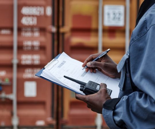 Close up of male worker wearing writing on clipboard in shipping docks with containers, copy space